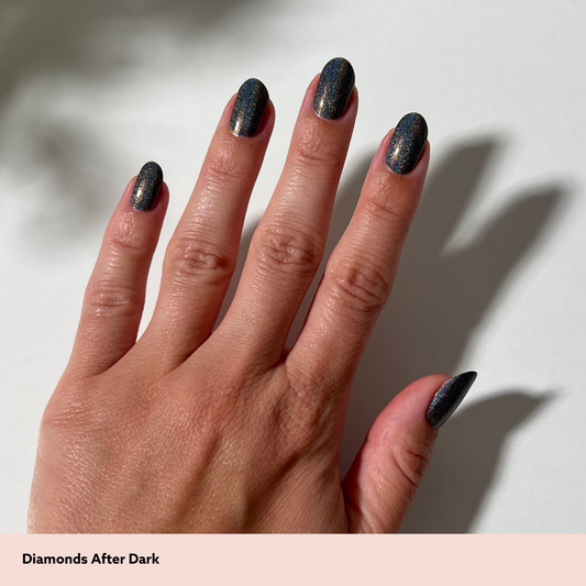 A close-up of a hand with long almond-shaped nails painted in a dark, shimmery charcoal nail polish that reflects blue and gold tones. The text 'Diamonds After Dark' appears at the bottom on a pale pink strip against a white background with soft shadows.