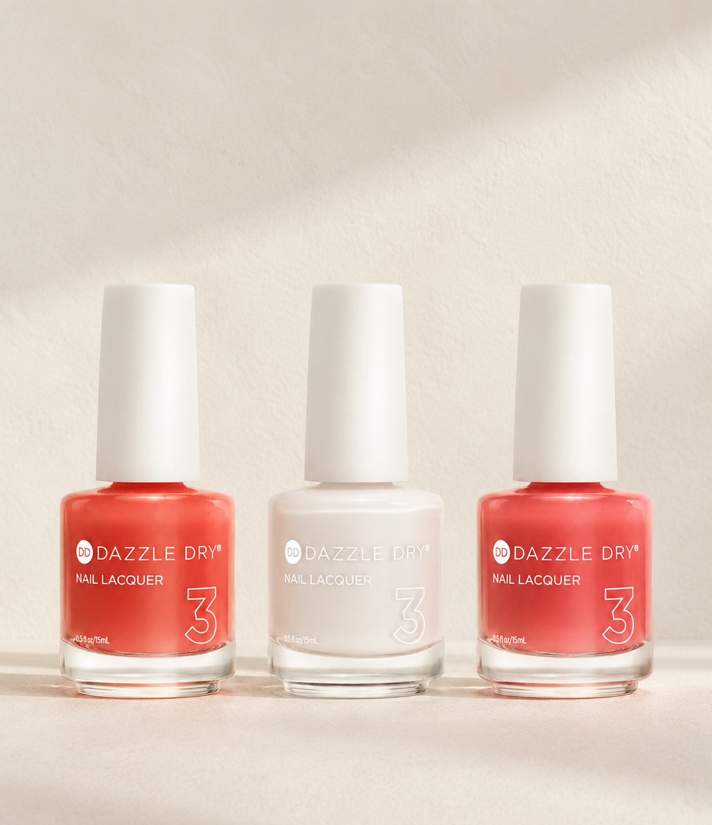 Three bottles of Dazzle Dry Nail Lacquer, numbers 3 in coral pink, white, and coral pink, arranged on a light beige textured background with soft diagonal shadows.