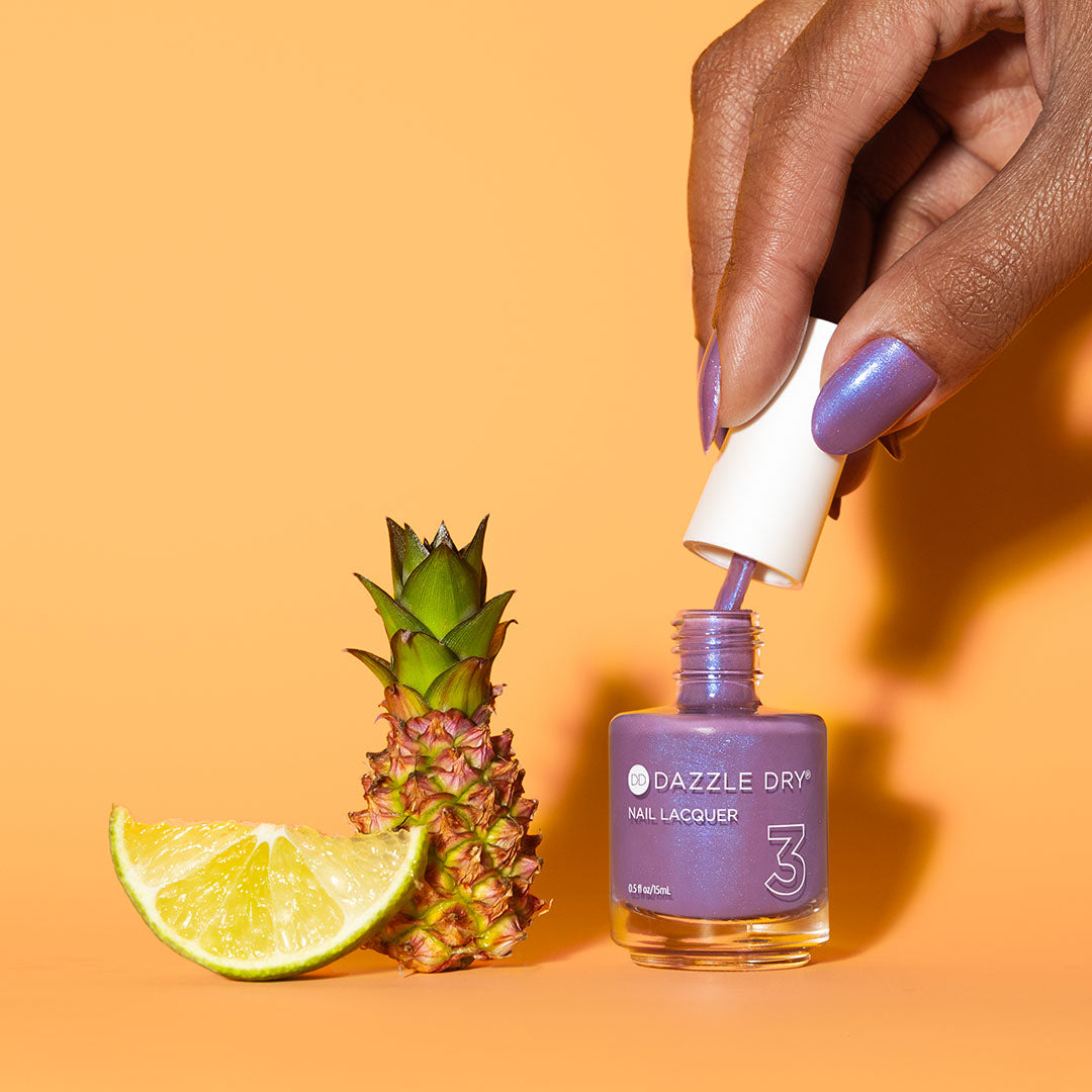Hand with Tropical Twilight painted on nails pulling lacquer brush out of the bottle next to a pineapple and lime slice