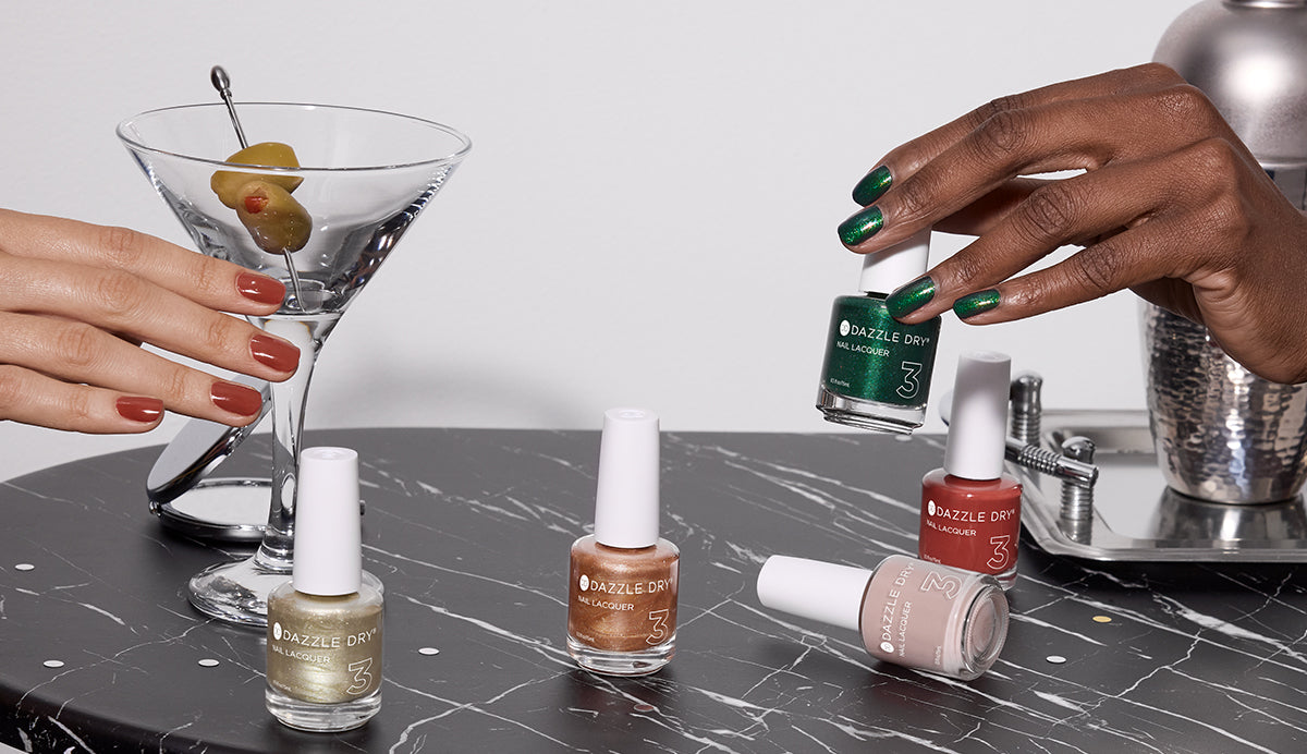 Dazzle Dry Afterparty Collection with red-painted hand reaching for a martini on a black-and-white veined marble table. Four lacquer bottles stand upright while Chiffon Moment lays down. Another hand with nails painted in Mistletoe holds the green color with a silver drink shaker behind.