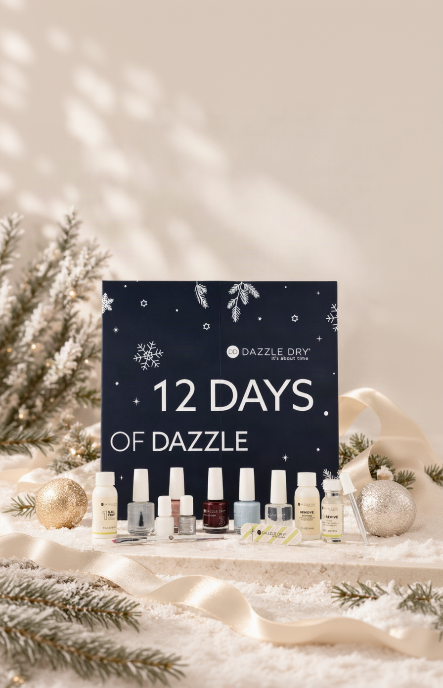 Dazzle Dry 12 Days of Dazzle nail polish advent box set, featuring a dark navy blue box with white snowflake designs and text, displayed with ten different nail polish bottles lined up in front on a festive, snowy holiday-themed background with gold ribbons and pine branches.