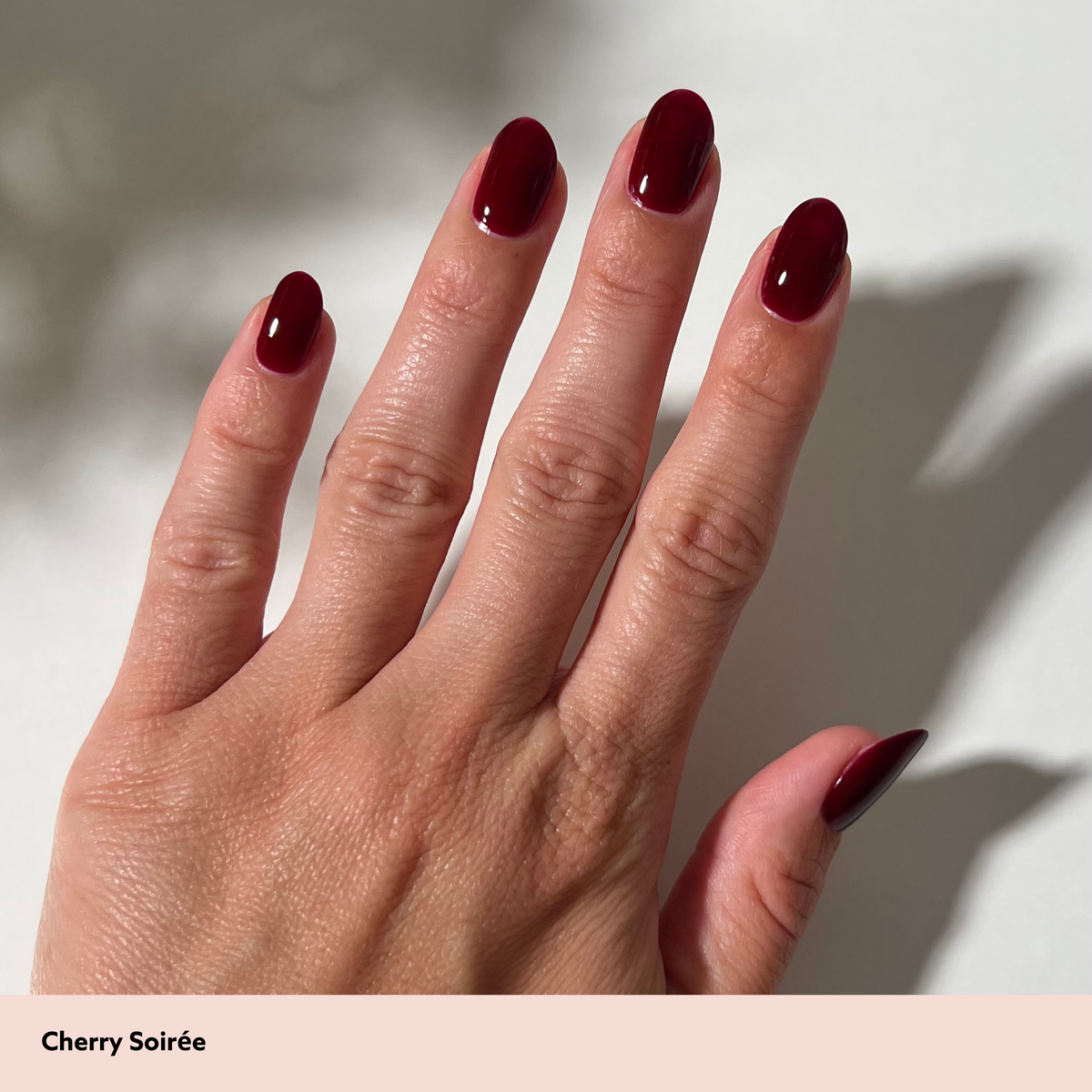 A close-up of a hand with long, almond-shaped nails painted in a glossy deep cherry red shade, labeled 'Cherry Soirée' on a pale pink strip at the bottom.