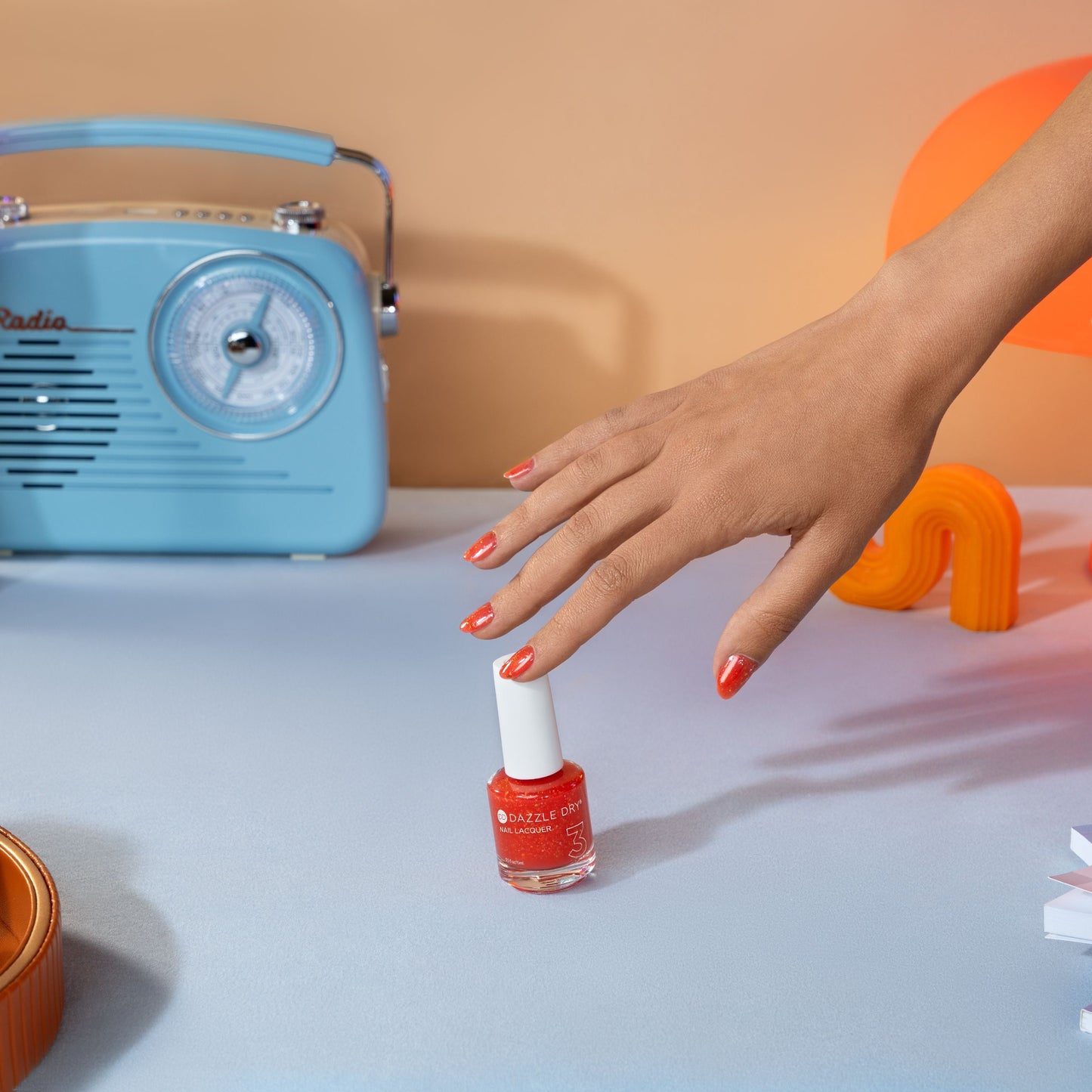 Hand with red nail polish holding a bottle of red nail polish next to a blue radio on a light surface.