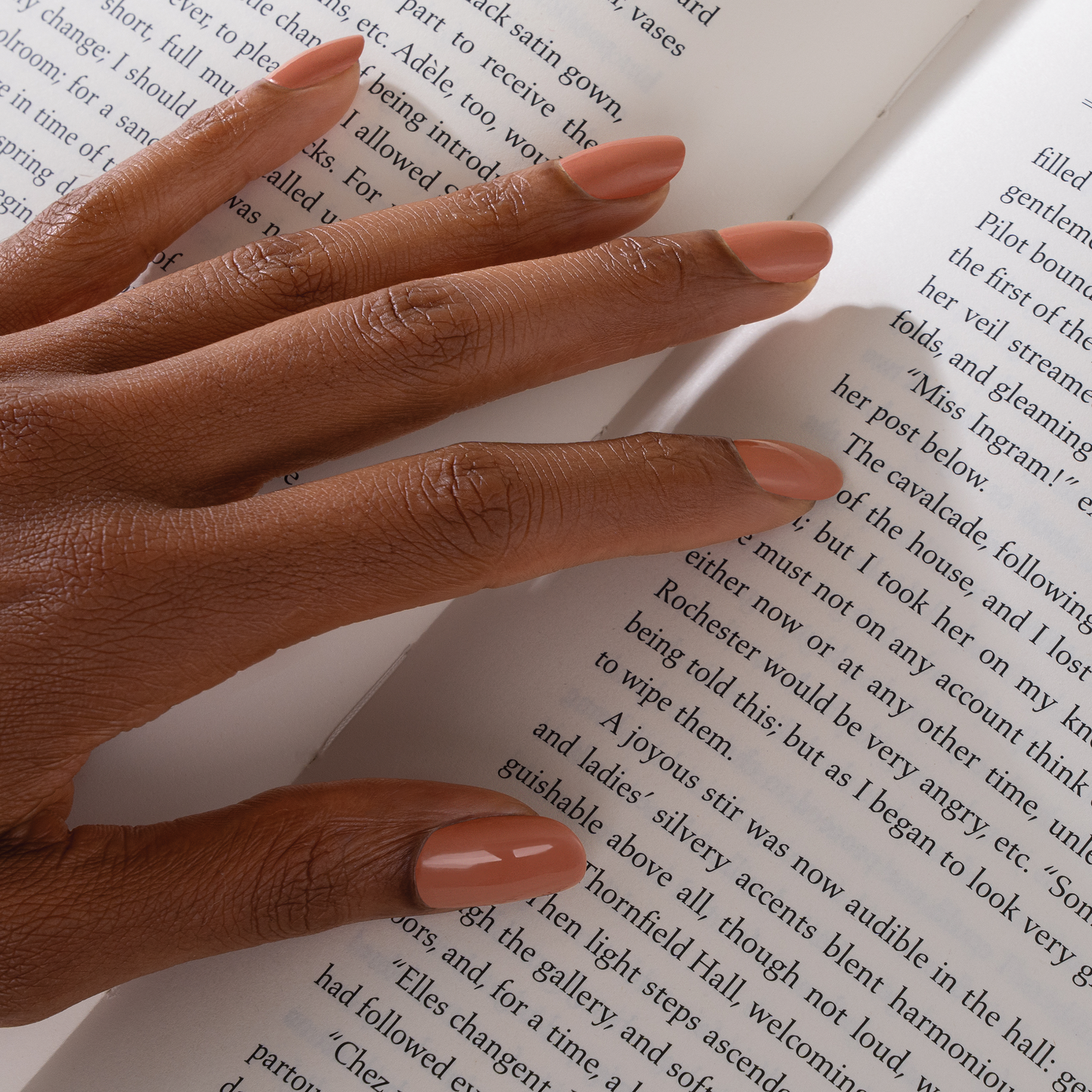 A close-up of a person's hand resting on an open book, showcasing a matte nude pink manicure.