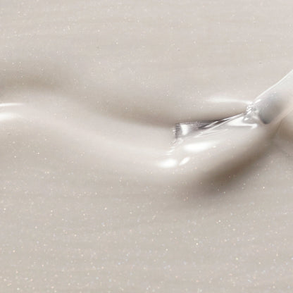 A close-up of a glossy, shimmering cream-colored cosmetic cream with fine silver glitter, swirled on a matching background with a small brush visible in the upper right.