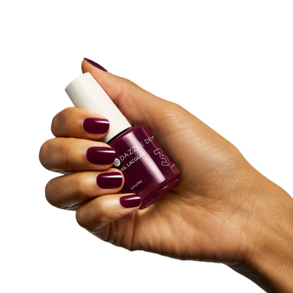 A hand with dark burgundy manicured nails holds a bottle of Darkberry Essentials nail polish against a solid white background.