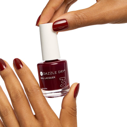 A bottle of Dazzle Dry Nail Lacquer in a deep burgundy shade, held by a hand with matching burgundy manicured nails against a solid white background.