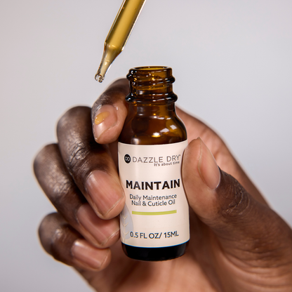 Hand holding a bottle of Dazzle Dry Maintain nail and cuticle oil with a dropper.