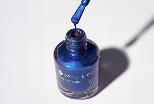 Dazzle Dry Fashion Week Blue nail lacquer with brush dripping polish back into the bottle, showcasing vibrant blue glitter color