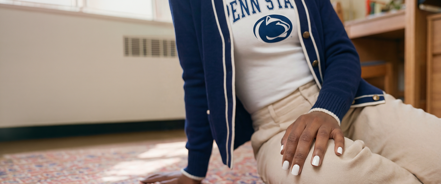 Woman sitting on a dorm room carpet leaning forward, wearing a Penn State blue cardigan with white stripes, a white T-shirt with the Penn State athletic logo, khaki slacks, and nails painted in Dazzle Dry White Lightning.