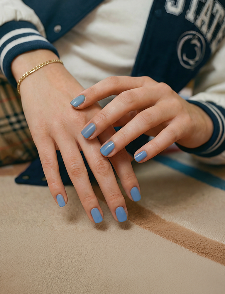 Woman leaning forward on a tan carpet wearing a Penn State varsity jacket with the Penn State athletic logo, dark blue and white sleeves with striped cuffs, a plaid dress over a white T-shirt, gold bralette and gold link bracelet, with light blue Dazzle Dry–painted nails.