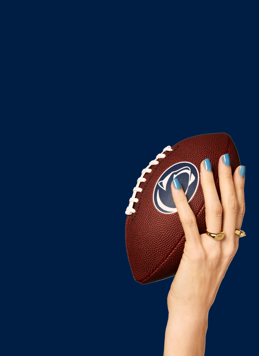 Hand holding a brown football featuring the Penn State Nittany Lion logo, with light blue nails painted in Dazzle Dry Varsity lacquer and Penn State gold rings.