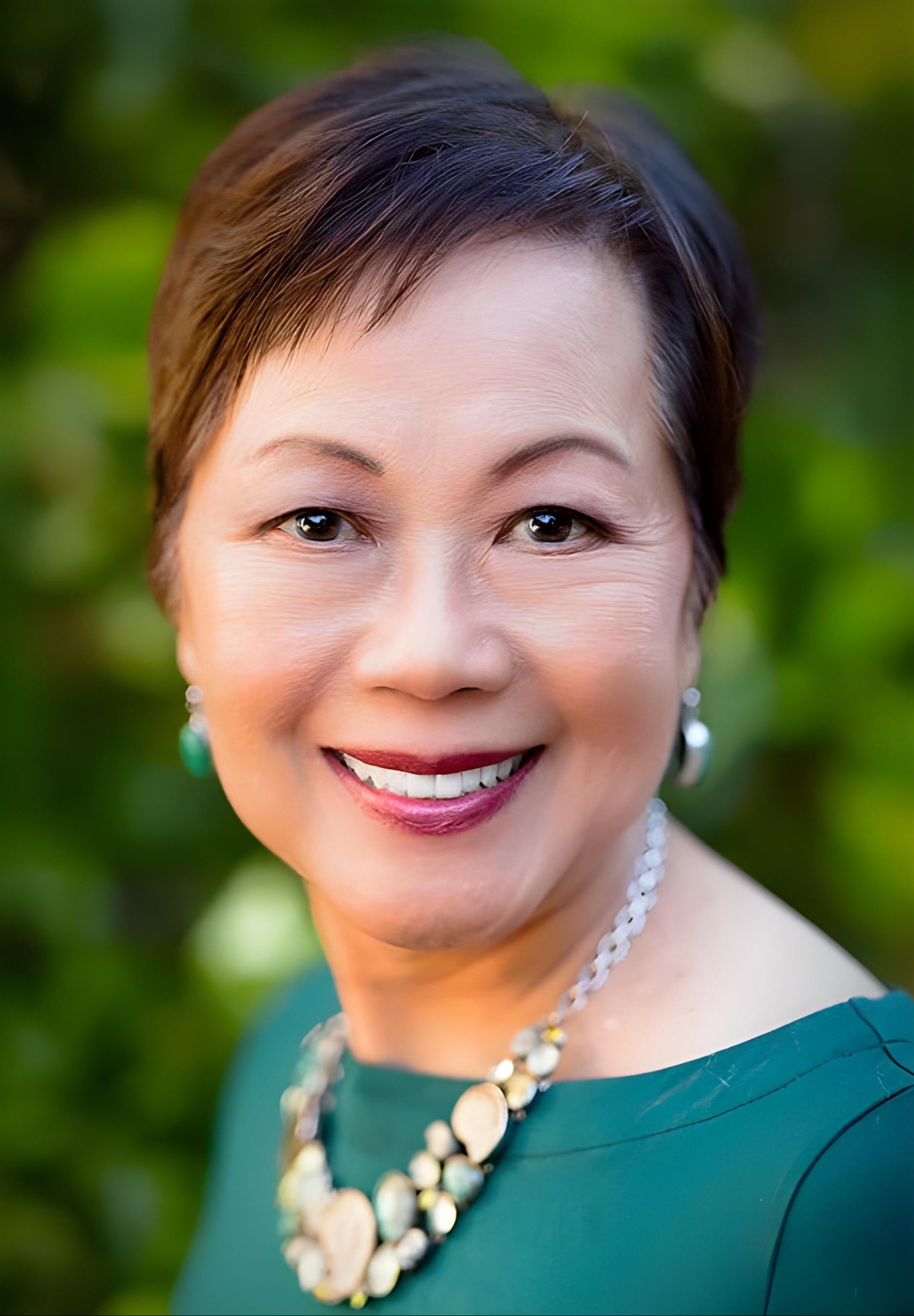 Dr. Vivian Valenty, inventor and founder of Dazzle Dry, smiling in a headshot. Short hair, red lipstick, green top, gold and silver necklace and earrings, in front of green bush background.
