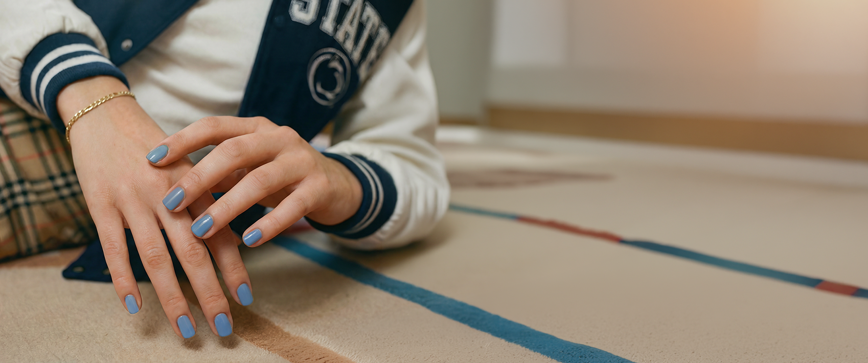 Woman leaning forward on a tan carpet wearing a Penn State varsity jacket with the Penn State athletic logo, dark blue and white sleeves with striped cuffs, a plaid dress over a white T-shirt, gold bralette and gold link bracelet, with light blue Dazzle Dry–painted nails.